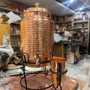 Puede incluir: Un dispensador de agua de cobre con una superficie texturizada, un grifo y una tapa con un pomo decorativo. Se encuentra sobre un soporte de metal negro con una taza de cobre a juego. El dispensador está diseñado para servir bebidas.