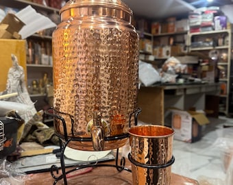 May include: A copper water dispenser with a textured surface, a spigot, and a lid with a decorative knob. It sits on a black metal stand with a matching copper cup. The dispenser is designed for serving beverages.