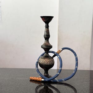 May include: An ornate black hookah with a blue hose and wooden mouthpiece sits on a dark surface. The hookah has intricate carvings and a decorative design. The blue hose is coiled, and the mouthpiece is light brown.