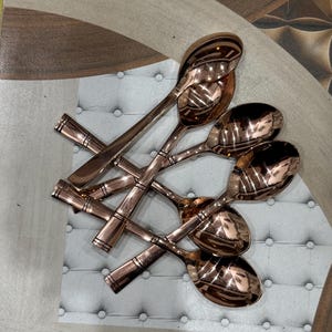 Puede incluir: Conjunto de seis cucharas de color cobre. Las cucharas tienen un acabado pulido que refleja la luz. Los mangos presentan un diseño texturizado. Las cucharas están dispuestas en un patrón entrecruzado.