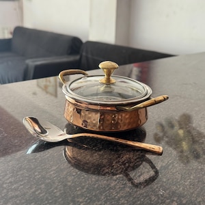 Puede incluir: Una olla de cocina de color cobre con tapa de cristal y pomo dorado, acompañada de una cuchara plateada. La olla tiene una textura martillada y asas pequeñas. La cuchara está colocada junto a la olla sobre una superficie oscura.