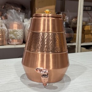 Könnte beinhalten: Ein Wasserspender aus Kupfer mit einem Wasserhahn und einem dekorativen Deckel. Der Spender hat eine gehämmerten Textur auf dem mittleren Abschnitt und einen goldfarbenen Edelstein oben. Das Design ist traditionell und handgefertigt.