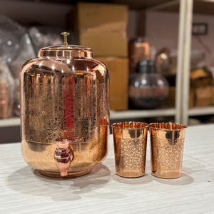 Puede incluir: Un dispensador de agua de cobre con grifo, tapa y dos vasos a juego. El dispensador y los vasos tienen un diseño floral grabado. Los artículos se exhiben sobre una superficie de color claro.