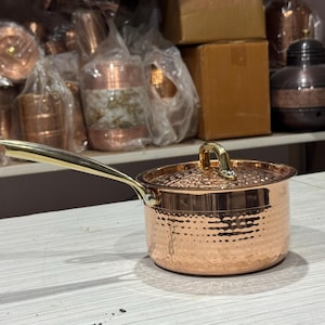 Puede incluir: Una cacerola de cobre con acabado martillado y asa y tapa doradas. La olla tiene aproximadamente 10 cm de diámetro y está sobre una superficie clara. Otros utensilios de cocina de cobre son visibles en el fondo.
