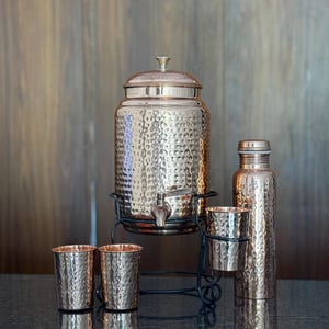 May include: A copper-colored hammered metal drink dispenser with a spigot, a matching water bottle, and several small cups. The dispenser sits on a black metal stand. The set is displayed on a dark surface.