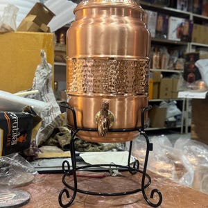 May include: A copper water dispenser with a spigot, lid, and decorative hammered band. The dispenser sits on a black wrought iron stand with scrolled feet. The dispenser is likely used for serving beverages.
