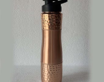 May include: A copper-colored water bottle with a black lid. The bottle has a hammered texture on the top and bottom sections, with a smooth, cylindrical middle section. The lid has a flip-top design.