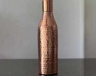 May include: A copper-colored water bottle with a hammered texture. The bottle has a cylindrical body that tapers slightly towards the top, and a flat, rectangular cap. The bottle is made of copper and has a shiny finish.