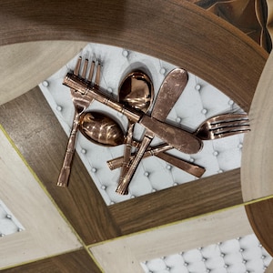 Op de afbeelding: Koperkleurige bestekset met vorken, lepels en messen. De gebruiksvoorwerpen zijn gerangschikt op een wit, gecapitonneerd oppervlak, met een decoratieve, geometrisch patroon achtergrond in bruin- en beigetinten.