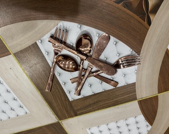 May include: Copper-colored flatware set including forks, spoons, and knives. The utensils are arranged on a white, tufted surface, with a decorative, geometric patterned background in shades of brown and beige.