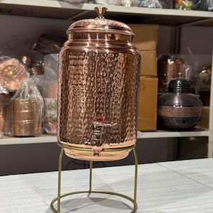 May include: A copper water dispenser with a hammered texture, a spigot, and a lid with a decorative finial. It sits on a gold-coloured metal stand. The dispenser is designed to hold liquids.