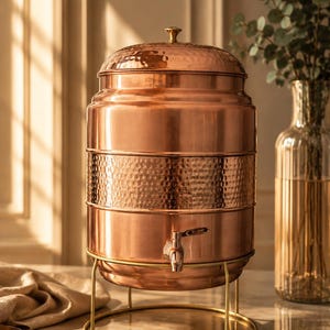 Puede incluir: Un dispensador de agua de cobre con textura martillada, un grifo y una tapa con un pequeño pomo. El dispensador se encuentra sobre un soporte de metal dorado. Un jarrón de cristal con vegetación se encuentra en el fondo.