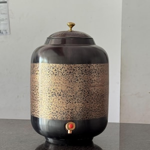 May include: A dark brown water dispenser with a gold-colored floral pattern. The dispenser has a lid with a gold knob and a red spigot. The dispenser is sitting on a dark surface.