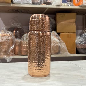 Könnte beinhalten: Eine Kupferwasserflasche mit strukturiertem, gehämmertem Design. Die Flasche hat einen passenden Deckel und eine zylindrische Form. Im Hintergrund sind weitere Kupferartikel zu sehen, was auf eine Sammlung von Kupferwaren hindeutet.