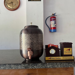 May include: A decorative copper water dispenser with an ornate floral pattern, a tap, and a lid. A clock, a fire extinguisher, and a sign that says "NO SMOKING" are visible in the background. A desk clock and a pen holder with the text "Ship2Gether" are also present.