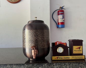 Può includere: Un distributore d'acqua color rame con un motivo floreale ornato, un rubinetto e un coperchio. Un orologio, un estintore e un cartello con la scritta "NO SMOKING" sono visibili sullo sfondo. Un orologio da scrivania e un portapenne con la scritta "Ship2Gether" sono anch'essi presenti.
