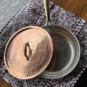 May include: A copper-colored frying pan with a lid, featuring a hammered texture. The pan has a silver interior and a gold-colored handle. The lid is partially open, revealing the interior. The pan rests on a patterned fabric.
