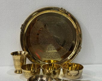 Puede incluir: Un juego de servir dorado que incluye una bandeja redonda grande, una taza alta, varios cuencos pequeños y una cuchara. La bandeja y la taza tienen diseños grabados ornamentados. Los cuencos y la cuchara son de diseño sencillo. El juego es probablemente para servir comida.