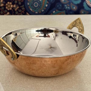 Könnte beinhalten: Eine Servierschüssel aus Kupfer und Edelstahl mit dekorativen goldfarbenen Griffen. Die Schüssel hat eine polierte Innenseite und eine strukturierte Außenseite. Die Griffe sind verziert und verleihen dem Design Eleganz. Geeignet für verschiedene Gerichte.