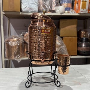 May include: A copper water dispenser with a hammered texture, a lid, and a spigot, on a black metal stand. A matching copper cup is attached. The dispenser has a "SAGA" sticker. The dispenser is likely to hold several litres.