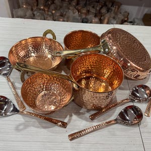May include: A selection of copper cookware and serving spoons. The copper pots and pans have a hammered texture and brass handles. The spoons are silver with hammered copper handles. The cookware includes a frying pan, saucepans, and bowls.