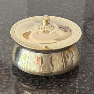 Op de afbeelding: Een glanzende, goudkleurige metalen container met een deksel. De container heeft een afgeronde body en een deksel met een kleine, decoratieve knop. Het oppervlak reflecteert licht, waardoor een heldere en gepolijste uitstraling ontstaat. De container wordt waarschijnlijk gebruikt voor opslag of weergave.