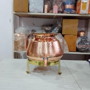 May include: A hammered copper water dispenser with a brass stand and tap. The dispenser has a wide, rounded body and a flared rim. The tap is blue. The dispenser is on a white table. Shelves in the background are filled with various items.