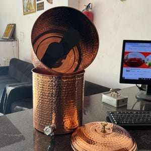 May include: A copper water dispenser with a hammered texture, a tap, and a lid. The dispenser is cylindrical and has a dark interior. The lid has a small, round handle. The dispenser is on a dark surface.