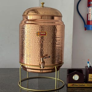 May include: A copper water dispenser with a hammered texture, a tap, and a lid with a decorative knob. The dispenser sits on a gold-coloured metal stand. The logo "SAGA" is visible on the front.