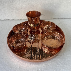 May include: A copper-coloured dining set with a hammered texture. The set includes a round tray, a tall cup, four small bowls, a spoon, and a fork. The items are arranged on a white surface.