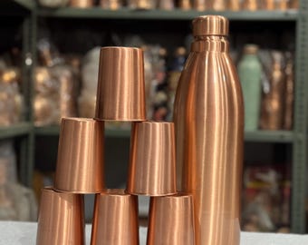 May include: A set of copper drinkware, including a tall water bottle and six small, conical cups. The items have a polished, metallic finish and are arranged on a white surface. The background shows shelves with more copper items.