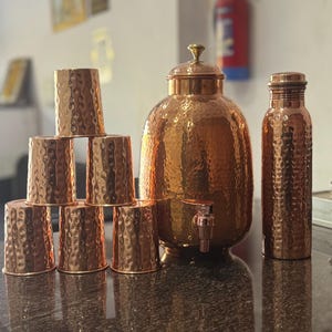 May include: A set of hammered copper drinkware, including a water dispenser with a tap, a tall water bottle, and seven small cups. The copper has a warm, reddish-brown hue. The cups are stacked in a pyramid shape.
