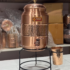 May include: A copper water dispenser with a hammered texture, a spigot, and a lid. The dispenser sits on a black metal stand with a matching copper cup. The SAGA logo is visible on the front of the dispenser.