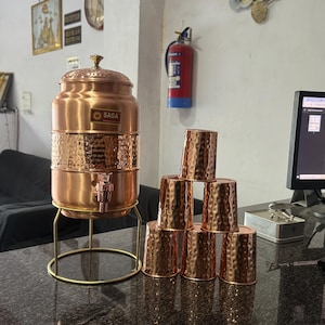 May include: A copper water dispenser with a hammered texture, a tap, and a lid, alongside six matching copper cups. The dispenser is on a gold-coloured stand. The word "SAGA" is visible on the dispenser.