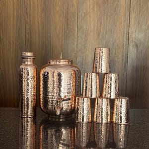 May include: A copper water set with a hammered texture. The set includes a tall bottle, a water dispenser with a spigot, and six matching drinking glasses. The copper pieces reflect the light, creating a warm, inviting aesthetic.