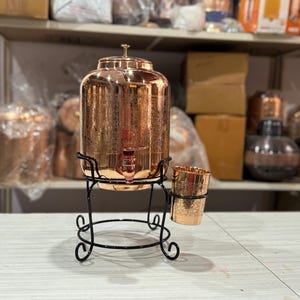 May include: A copper-colored water dispenser with a spigot, lid, and a matching cup holder. The dispenser sits on a black metal stand with a circular base and decorative swirls. The background includes shelves with various items.