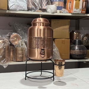 May include: A copper water dispenser with a hammered texture, a tap, and a lid, set on a black metal stand. A matching copper cup sits beside it. Other copper items are visible on shelves in the background.