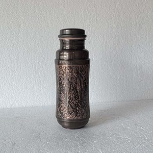 Peut inclure: Une gourde cylindrique marron foncé avec un bouchon à vis. La bouteille présente un motif texturé en forme d'arbre sur la partie centrale. Le design est moderne et élégant, idéal pour l'eau ou d'autres boissons.