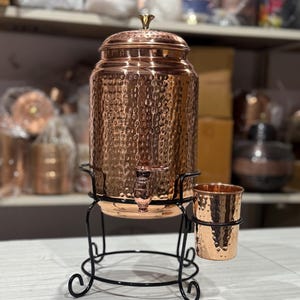 Peut inclure: Un distributeur d'eau en cuivre avec une texture martelée, un couvercle et un robinet. Il repose sur un support en métal noir avec un porte-gobelet assorti et deux gobelets en cuivre. Conçu pour servir des boissons.