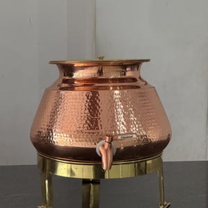 May include: A copper-coloured hammered metal beverage dispenser with a tap, set on a brass-coloured stand. The dispenser has a lid and a label that reads "Classinox". The stand has three legs.