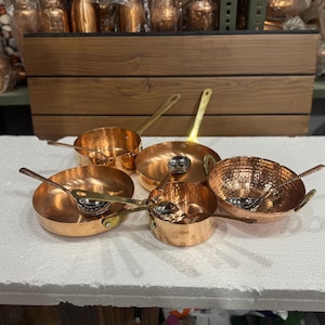 May include: A collection of copper cookware, including small pans and bowls, with brass handles and serving spoons. The copper has a shiny, reflective surface. The cookware is arranged on a white surface.