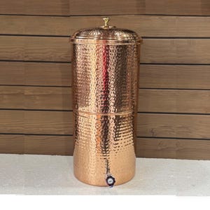 Op de afbeelding: Een koperen waterfilter met een gehamerde textuur. De cilindrische container heeft een deksel met een kleine knop en een kraan aan de basis. Het filter is ontworpen om water te zuiveren en heeft een decoratieve uitstraling.