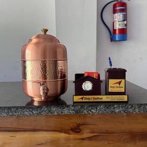 May include: A copper water dispenser with a hammered texture, a tap, and a gold-coloured knob. A small clock and business card holder with the text "Ship2Gether" are also visible. A pen holder and a fire extinguisher are in the background.