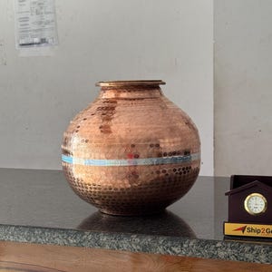 Könnte beinhalten: Eine kupferfarbene, runde Vase mit breitem Körper und schmalem Hals. Die Vase hat eine strukturierte Oberfläche mit einem Band farbiger Akzente in der Mitte. Die Vase steht auf einer dunklen Oberfläche.