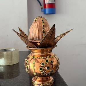 Op de afbeelding: Een koperkleurige decoratieve pot met een deksel en bladvormige accenten. De pot is versierd met groene en rode edelstenen en goudkleurige details. De pot staat op een donker oppervlak.