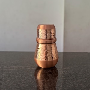 Puede incluir: Un recipiente de color cobre con acabado martillado y tapa a juego. El recipiente tiene una base redondeada y una parte superior cilíndrica. La superficie del recipiente y la tapa tienen un patrón texturizado y con hoyuelos. Se encuentra sobre una superficie oscura y moteada.