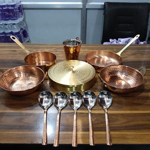 以下が含まれることがあります： 木製の表面に配置された銅製の調理器具とサービングスプーンのコレクション。 セットには、ハンマー加工された銅製の鍋、ボウル、バケツ、銅製のハンドルが付いた磨かれたシルバースプーンが含まれています。 ふた付きの真鍮製の容器も含まれています。