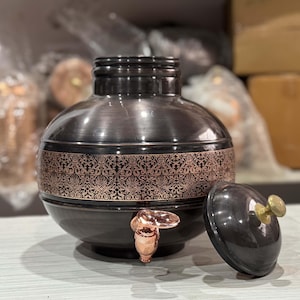 May include: A dark metallic water dispenser with a copper-colored floral design band. The dispenser has a spigot and a lid with a brass knob. The lid is off to the side. The dispenser is on a light-colored surface.