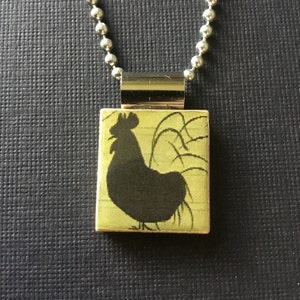 May include: A silver chain necklace with a rectangular pendant featuring a black silhouette of a rooster on a green background.