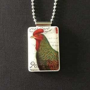 May include: A silver chain necklace with a rectangular pendant featuring a vintage postcard image of a rooster with a red comb and green and brown feathers. The postcard says "Post Card" and "Po" at the bottom.